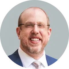 Smiling man in a suit with glasses, beard, and short hair against a plain background.
